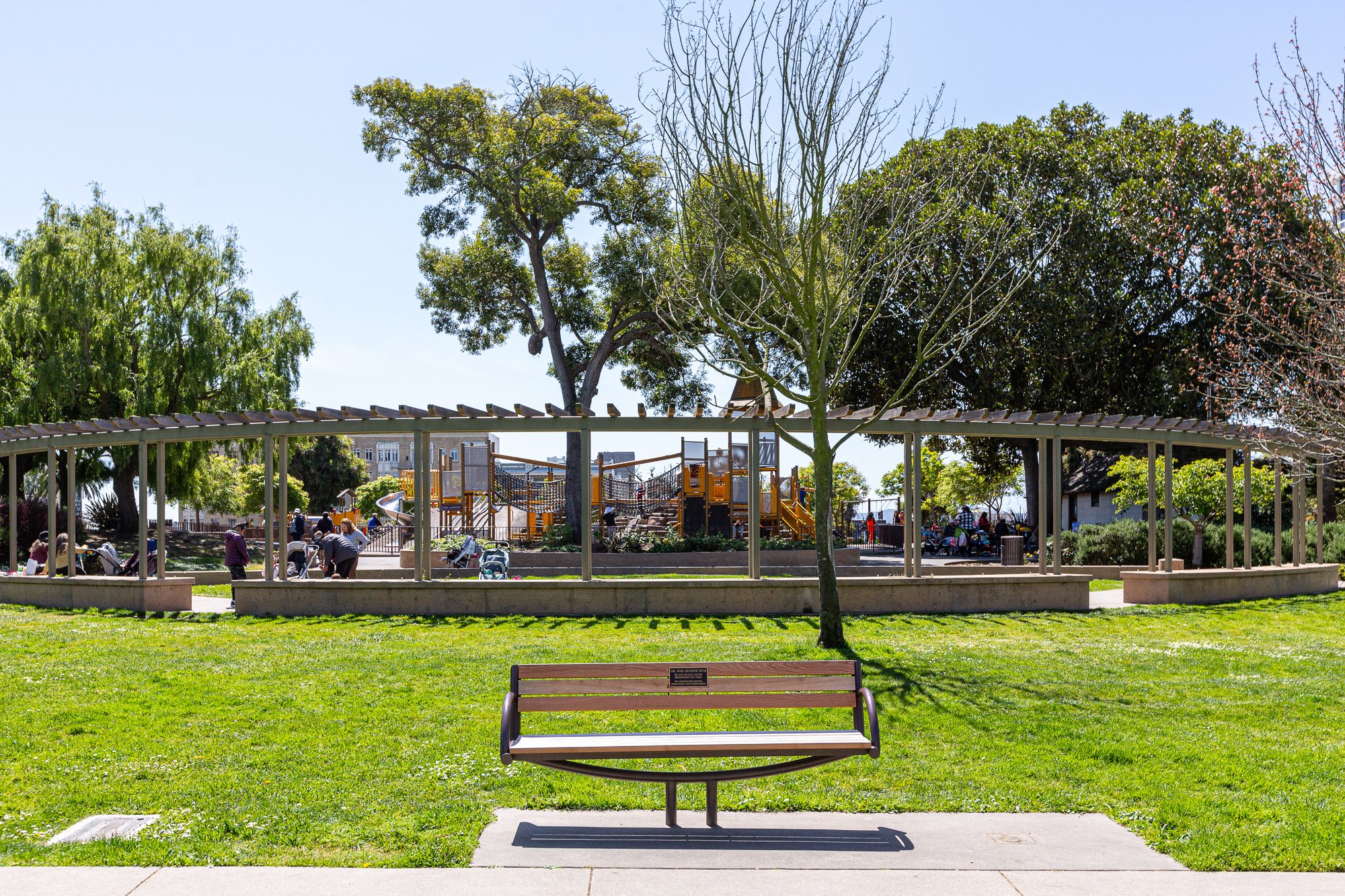 Park with grassy area, benches, trees, playground with slides and climbing structures, and people sitting and walking around.