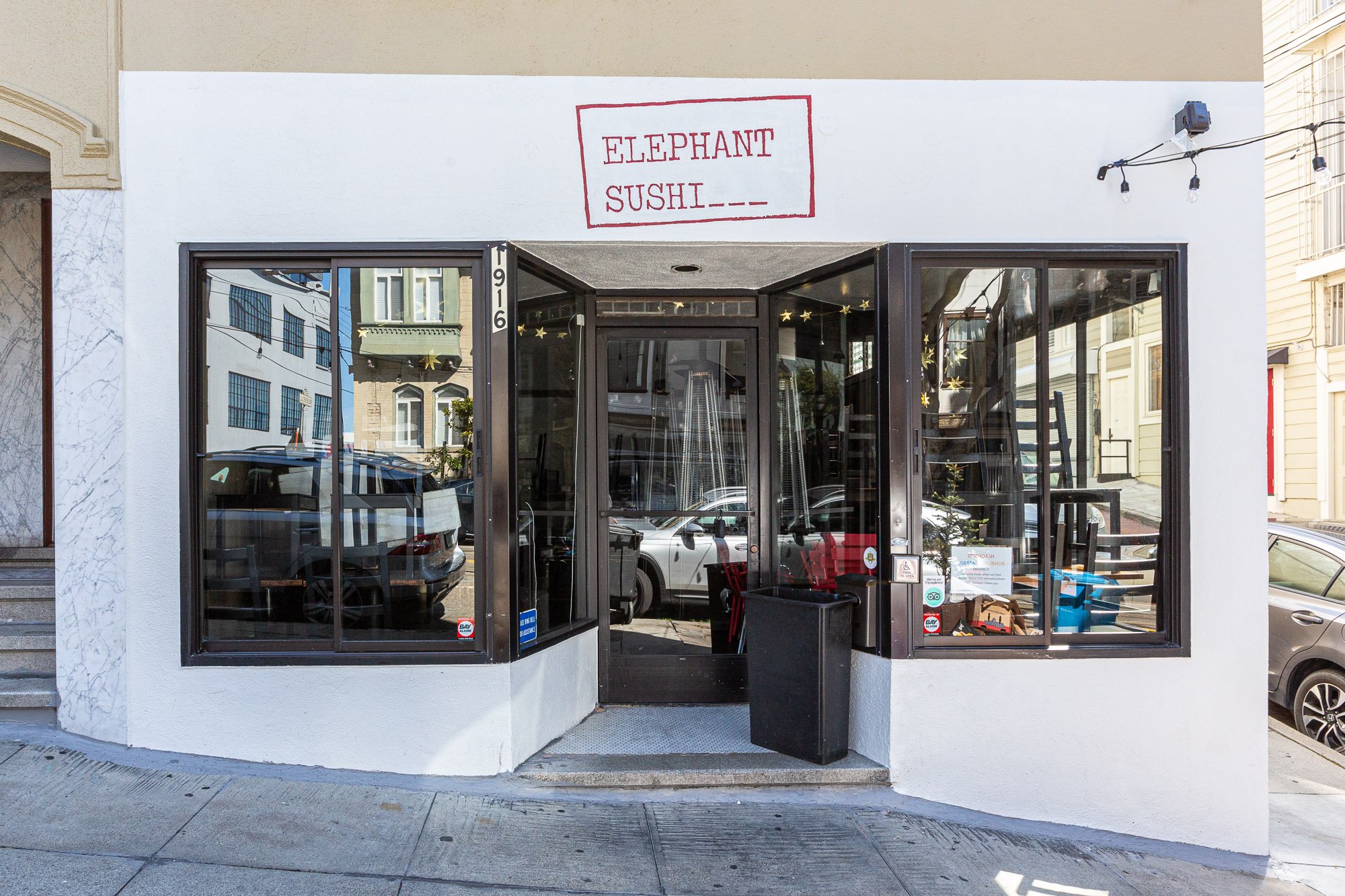 Front view of a small sushi restaurant named Elephant Sushi with black-framed glass windows and an entrance door, decorative star lights inside, and a handwritten sign on the wall above the door.