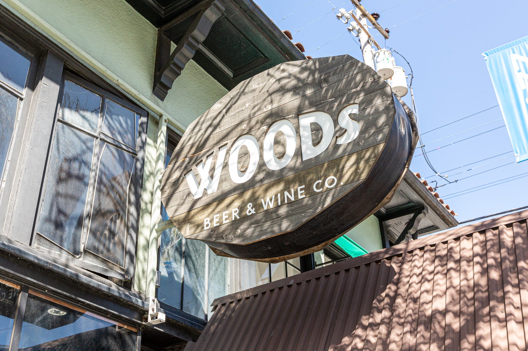 Wooden sign for Woops Beer & Wine Co. hangs outside a building with windows and a brown metal roof, against a clear blue sky.