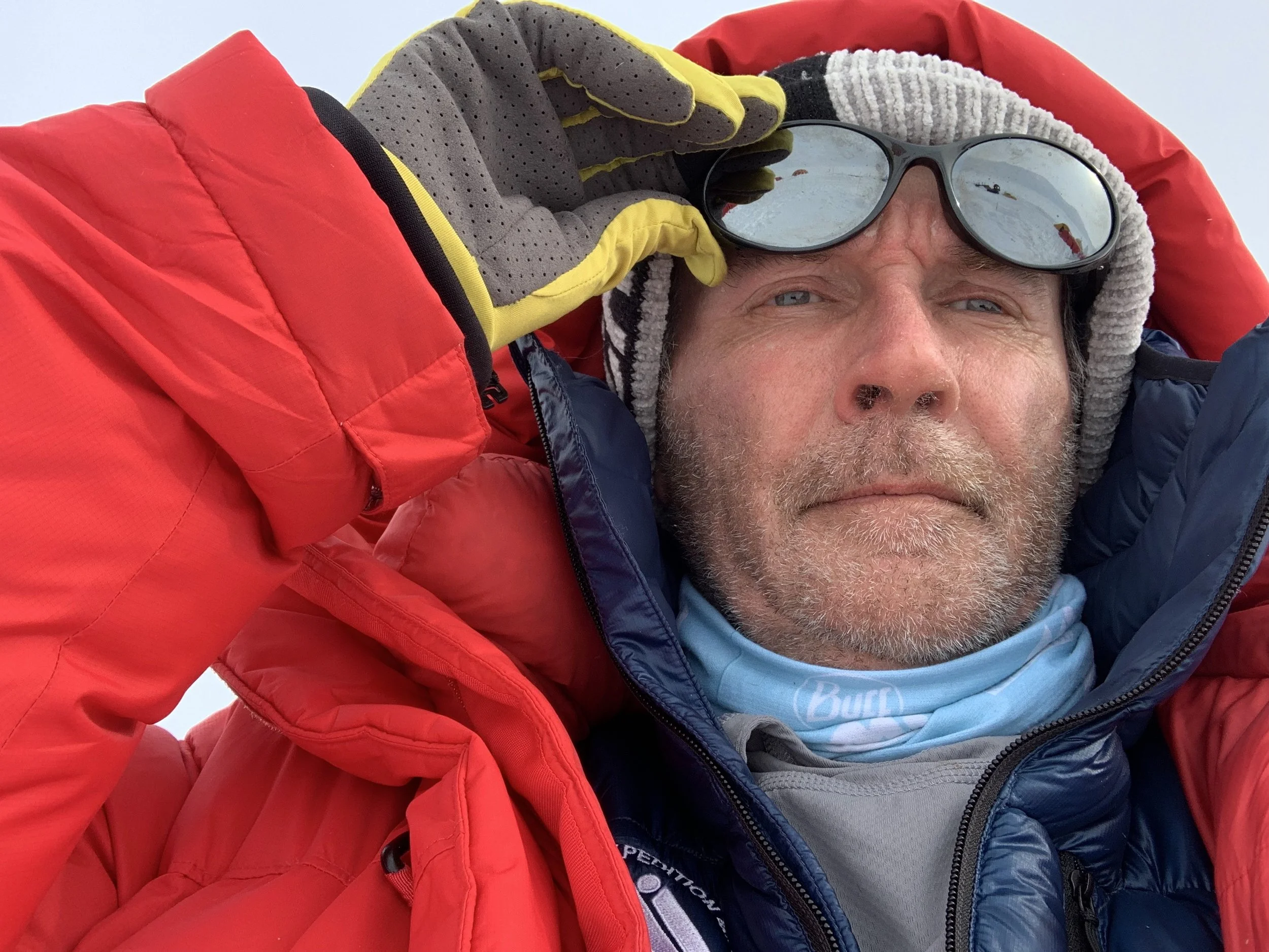 A man in winter gear with a red, blue, and gray jacket, gray hat, sunglasses, and a neck gaiter, taking a selfie outdoors in a cold environment.
