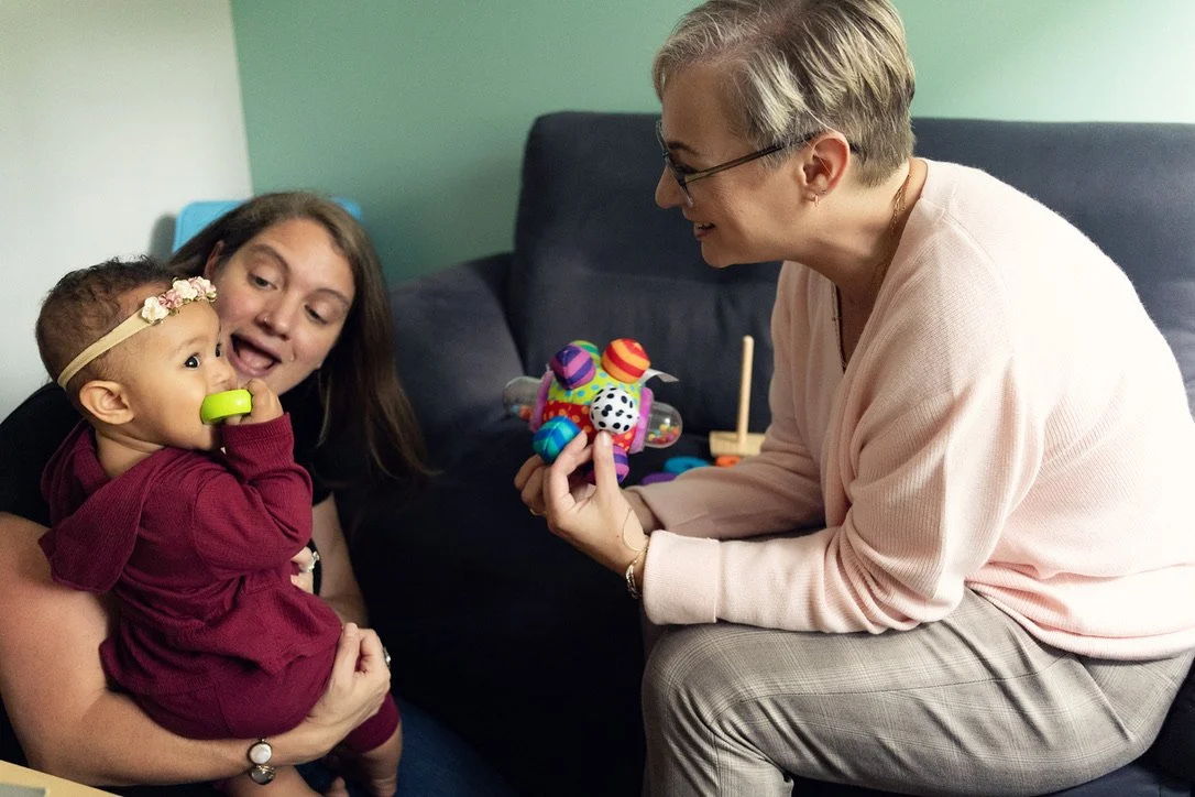 A woman holding a baby with a headband, smiling at an older woman who is showing her a colorful toy, in a cozy living room.
