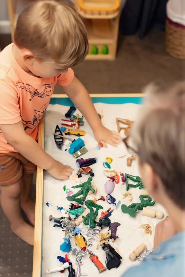 Two children playing with a variety of small plastic toys on a white surface, in a room with wooden shelves and a basket.