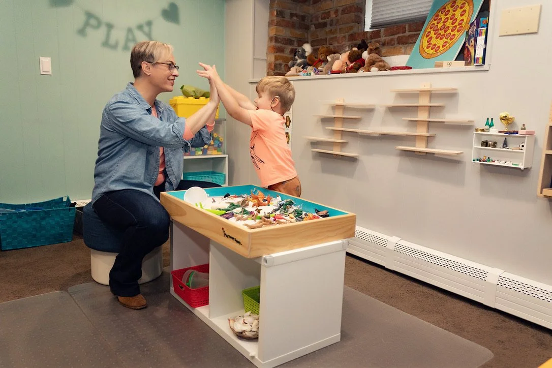 A woman and a young boy are playing together in a playroom, giving each other a high five. The room has toys, shelves, and a large pizza picture on the wall.