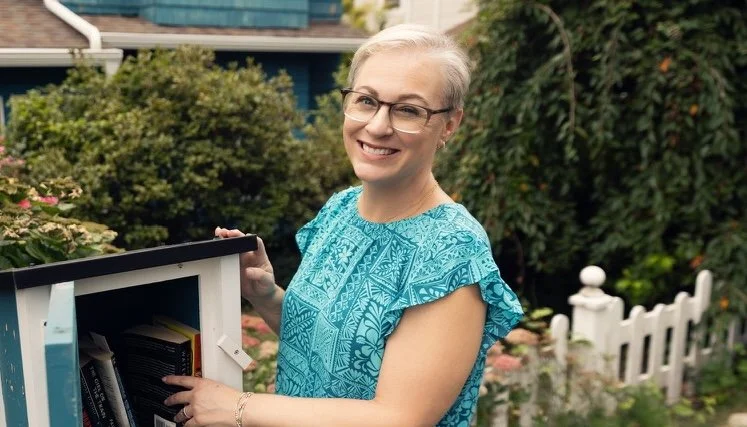 A woman with glasses in a blue patterned top standing outdoors near a bookshelf, smiling.