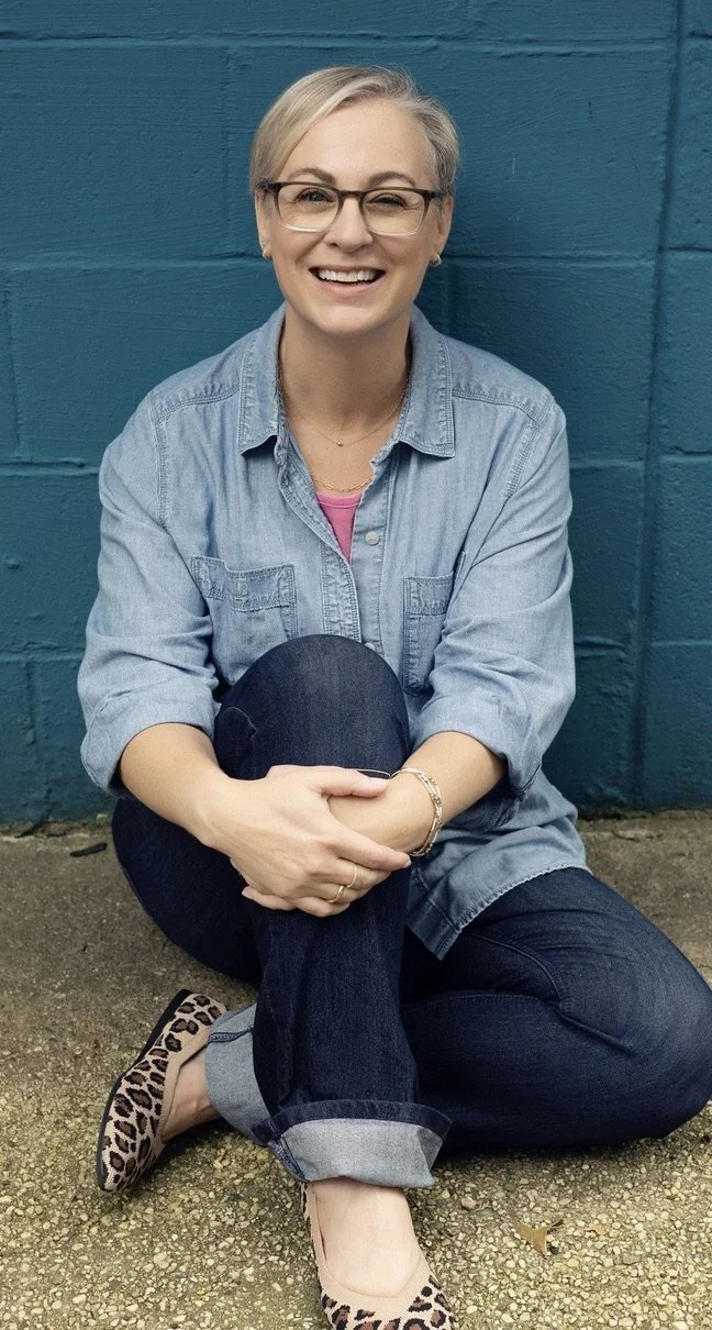 A smiling woman with short blonde hair, glasses, and a denim shirt sitting on the ground with legs crossed, in front of a blue wall.