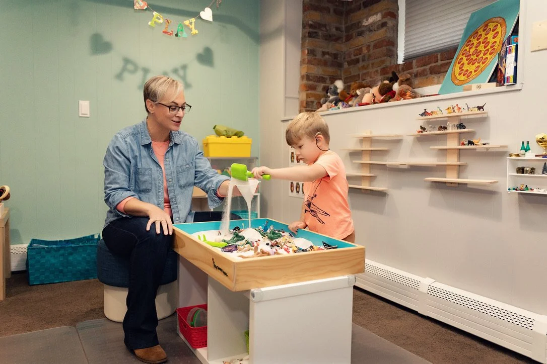 A woman and a young boy playing with toy dinosaurs and figurines at a sensory table in a playroom with light green and brick walls, shelves with toys, and a banner that says 'PLAY' on the wall.