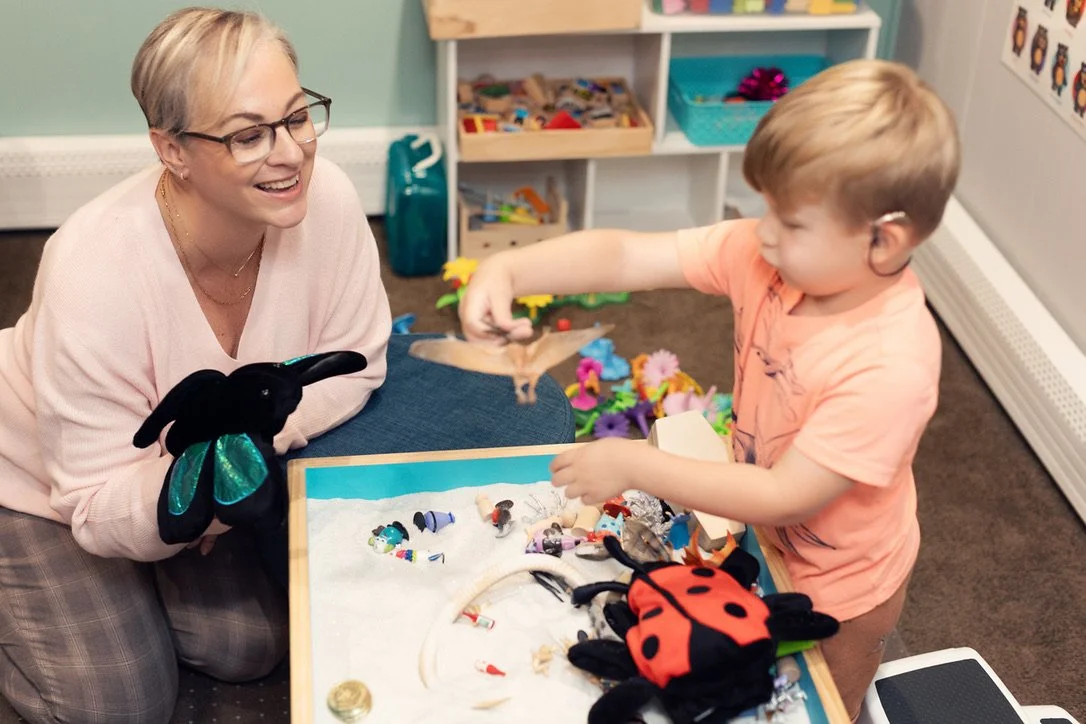 A woman and a young boy are playing with plush toys of a ladybug, a butterfly, and other animals at a table with a sand-filled display. The woman is smiling and wearing glasses, while the boy is reaching out with his hand. The background shows shelves with toys and colorful decorations.