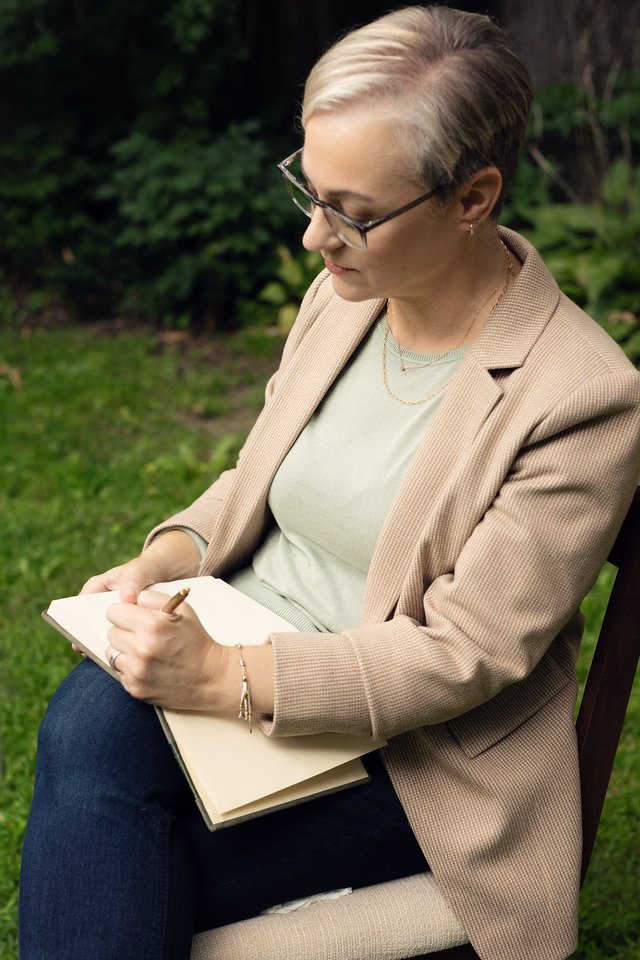 A woman with short blonde hair, glasses, and a beige blazer sitting on a wooden chair outdoors, writing in a notebook.