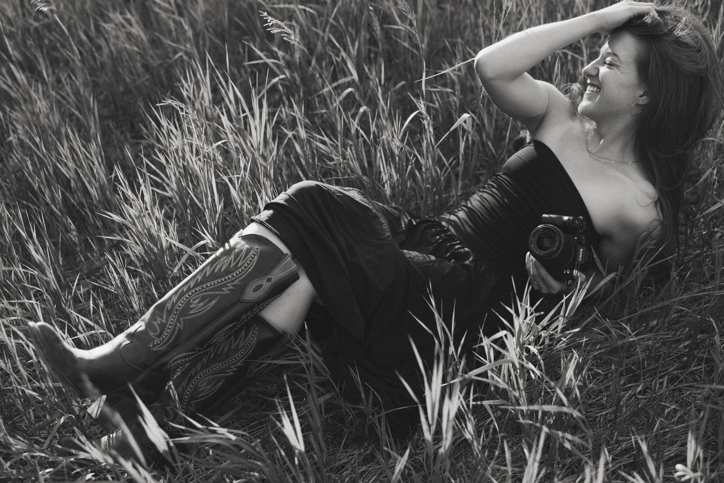 A smiling woman lying on her side in a grassy field, holding a camera in her right hand, with her left hand running through her hair.