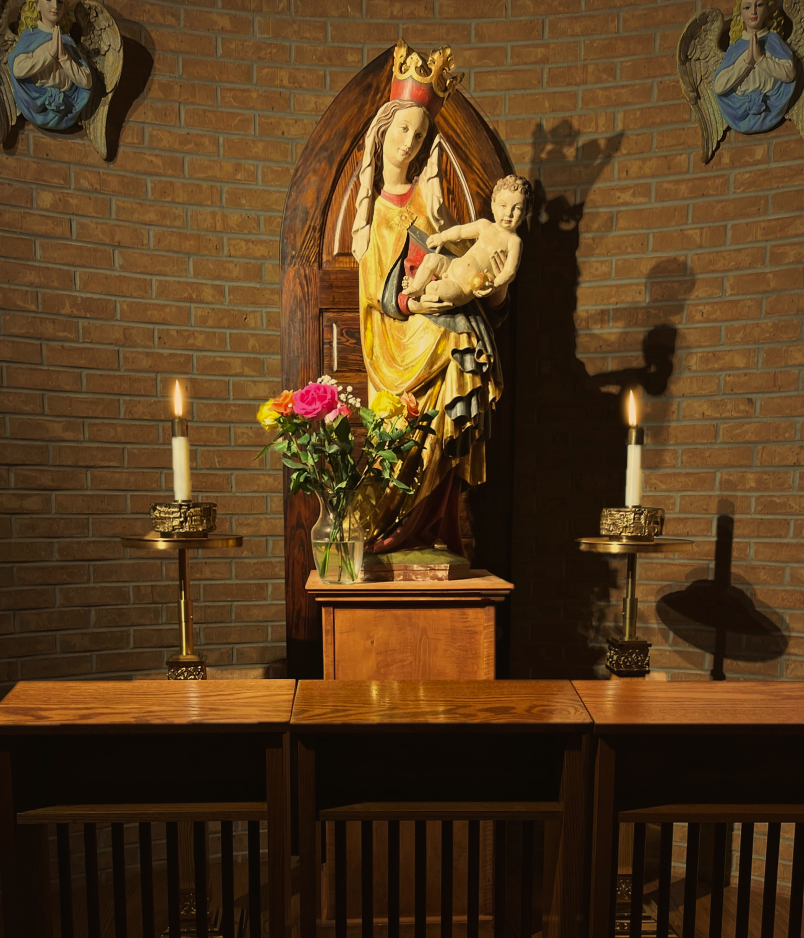A religious altar with a wooden statue of the Virgin Mary holding baby Jesus, flanked by two candles and a vase of multicolored roses, against a brick wall.