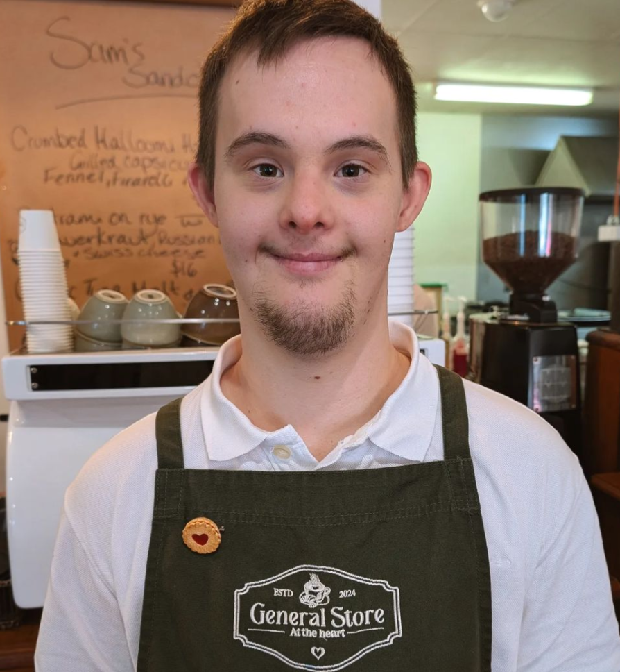 Young man wearing a white shirt and a black apron with the logo "General Store" stands inside a coffee shop. There are coffee cups and a coffee grinder in the background.