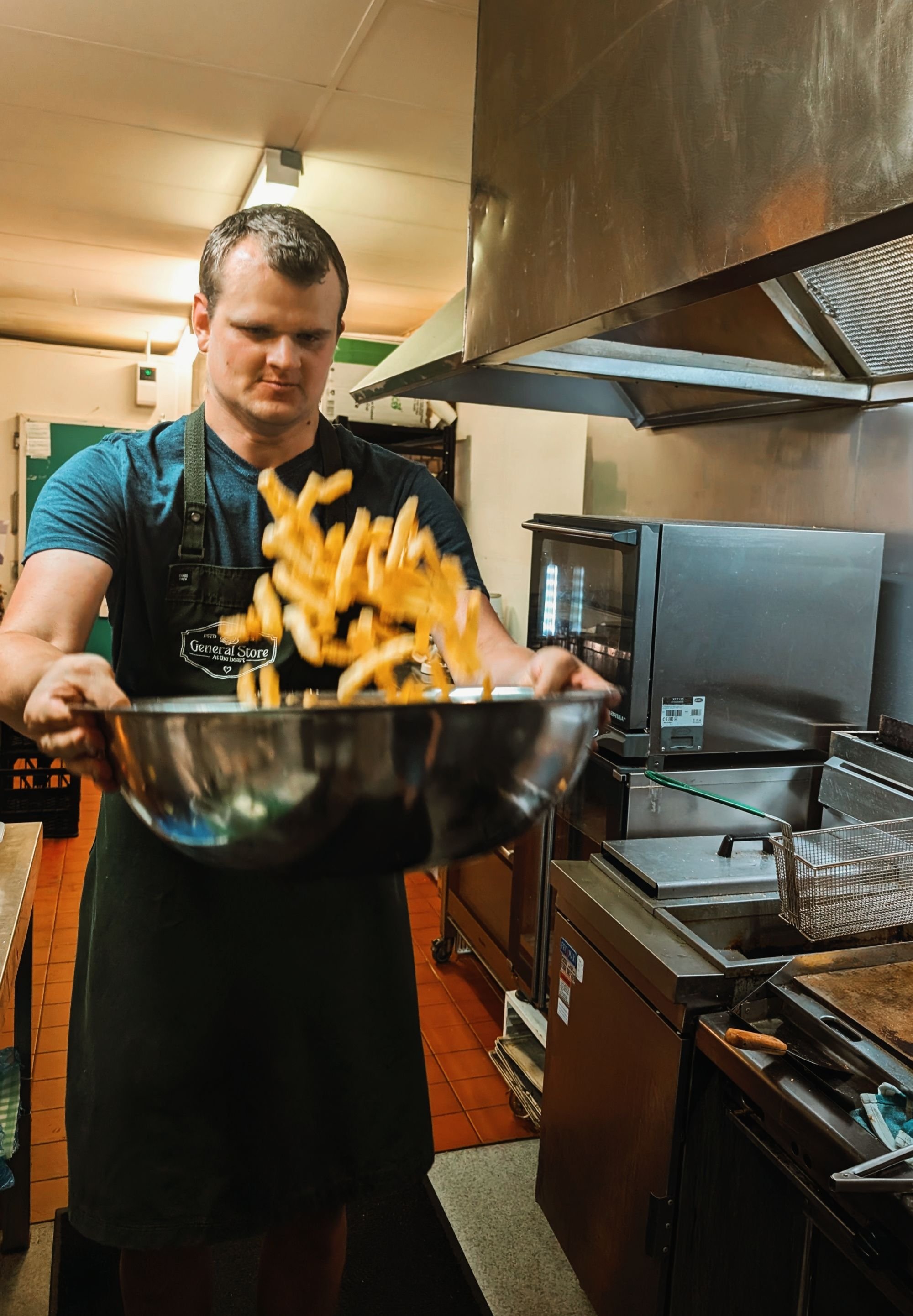 A young man in a black apron tossing french fries in a large metal bowl in a commercial kitchen.