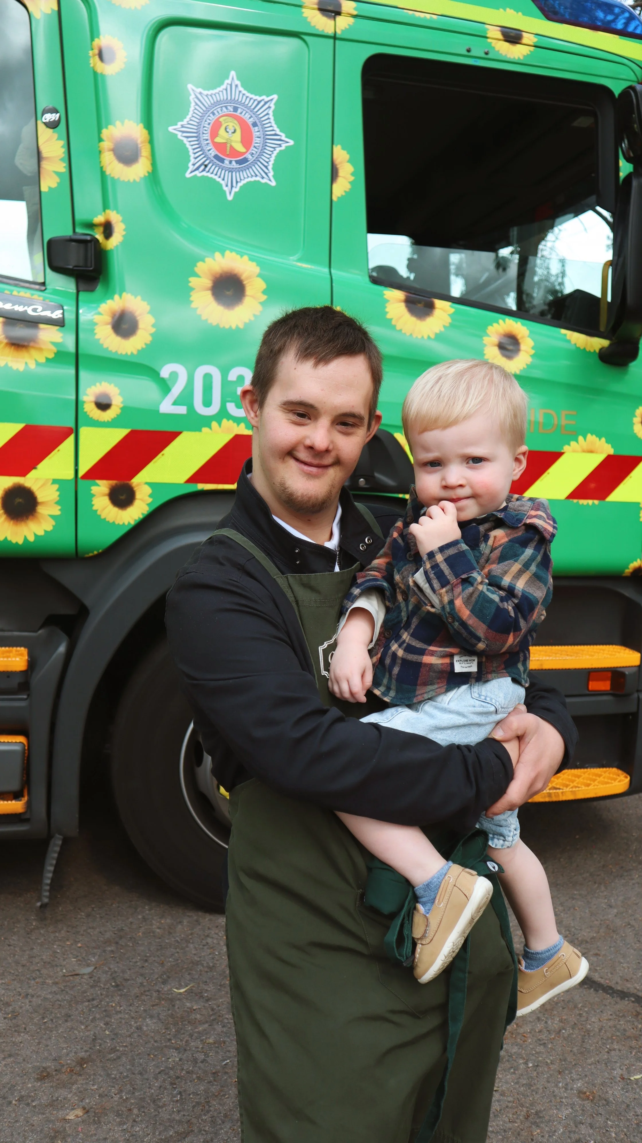 A firefighter holding a young boy in front of a colorful fire truck decorated with sunflowers.