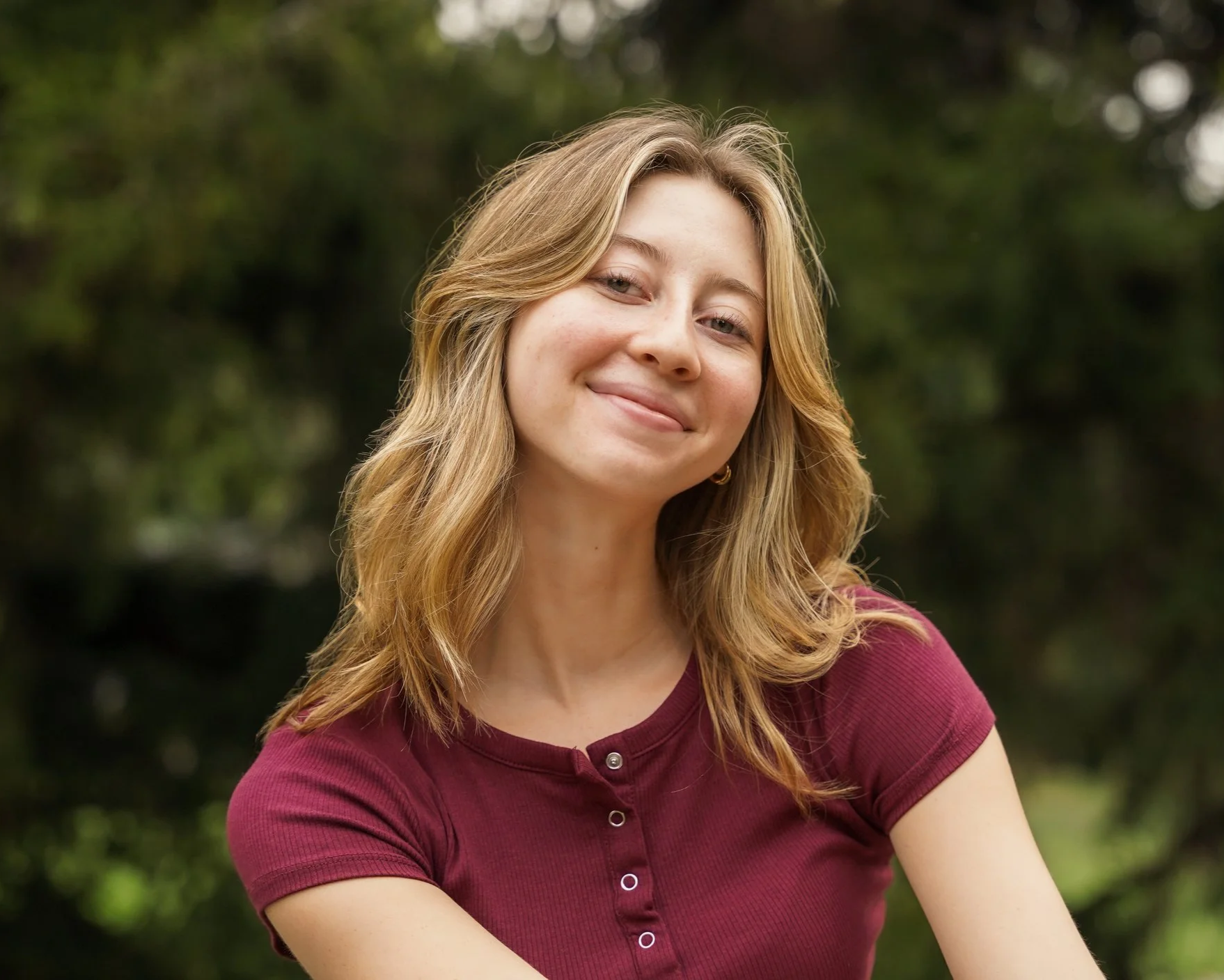 A young woman with shoulder-length, wavy blonde hair, smiling and looking at the camera, outdoors with greenery in the background, wearing a maroon short-sleeve top.