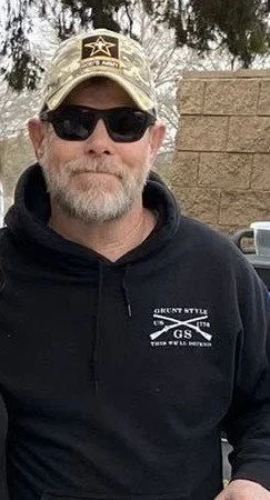 Man wearing a camouflage cap with a Texas flag patch and a star badge, dark sunglasses, and a gray shirt, outdoors with green foliage in the background.