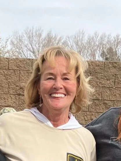 A smiling woman with blonde hair standing outdoors in front of a brick wall and leafless trees.