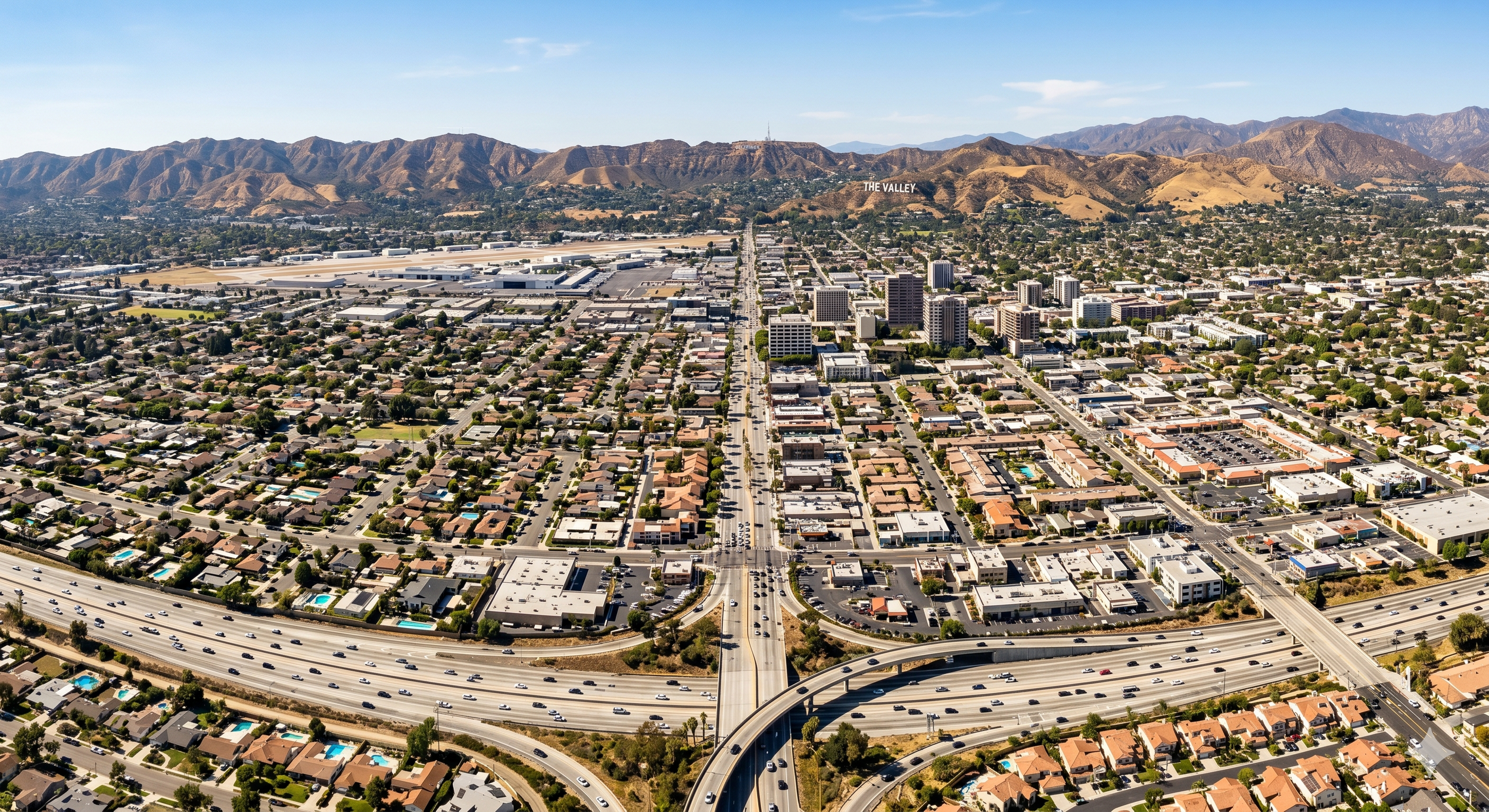 An aerial view of a city with residential neighborhoods, commercial buildings, and highways, with mountains in the background.