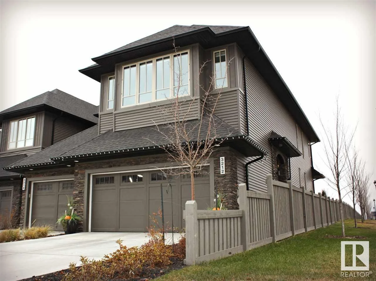Modern two-story house with a gray exterior, large windows, a three-car garage, and a wooden fence in front. There is a small leafless tree and some plants near the driveway.
