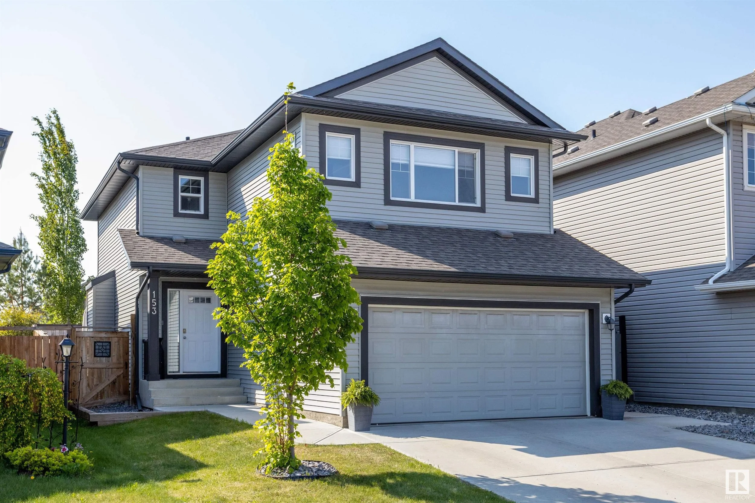 A modern two-story house with gray siding, a gabled roof, a two-car garage, and a front yard with a small tree and plants.