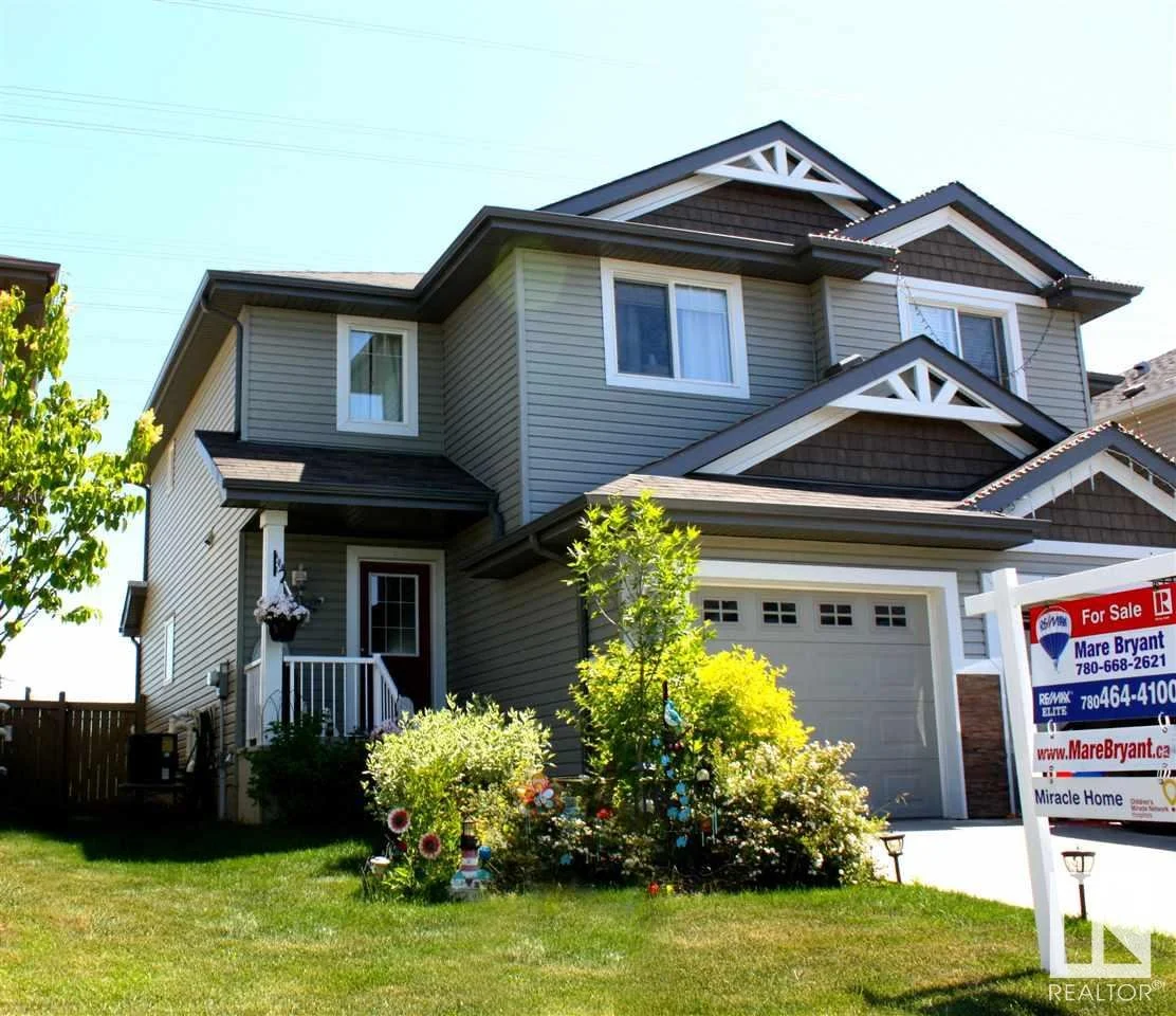 A modern three-story house with beige siding and brown shingles, a white garage door, and a lawn with colorful outdoor ornaments. A "For Sale" sign is displayed in the yard.