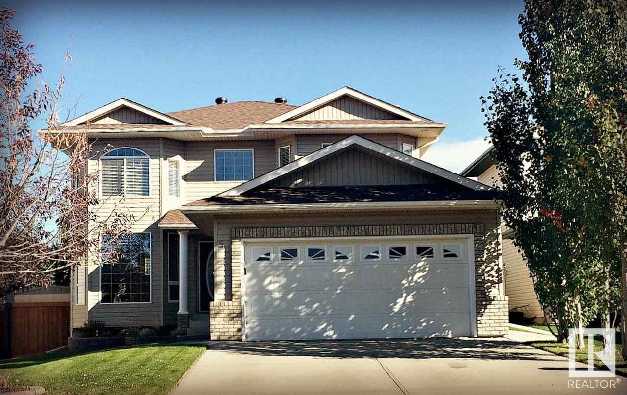 A two-story house with beige siding, a brown shingle roof, white garage door, and front porch with brick columns. Trees with autumn foliage are on either side of the house, and the lawn is green.