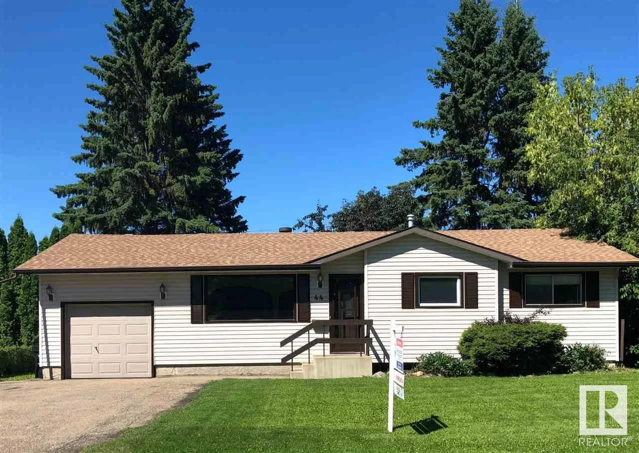 Single-story house with white siding, brown shutters, a small front porch, attached garage, and a well-maintained lawn with trees in the background under a clear blue sky.