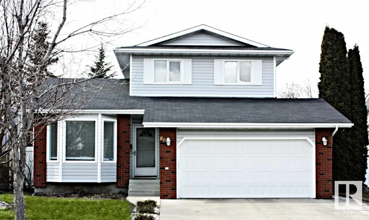 A two-story house with red brick and light gray siding, white garage door, front door, and window shutters, with bare tree and a small lawn.