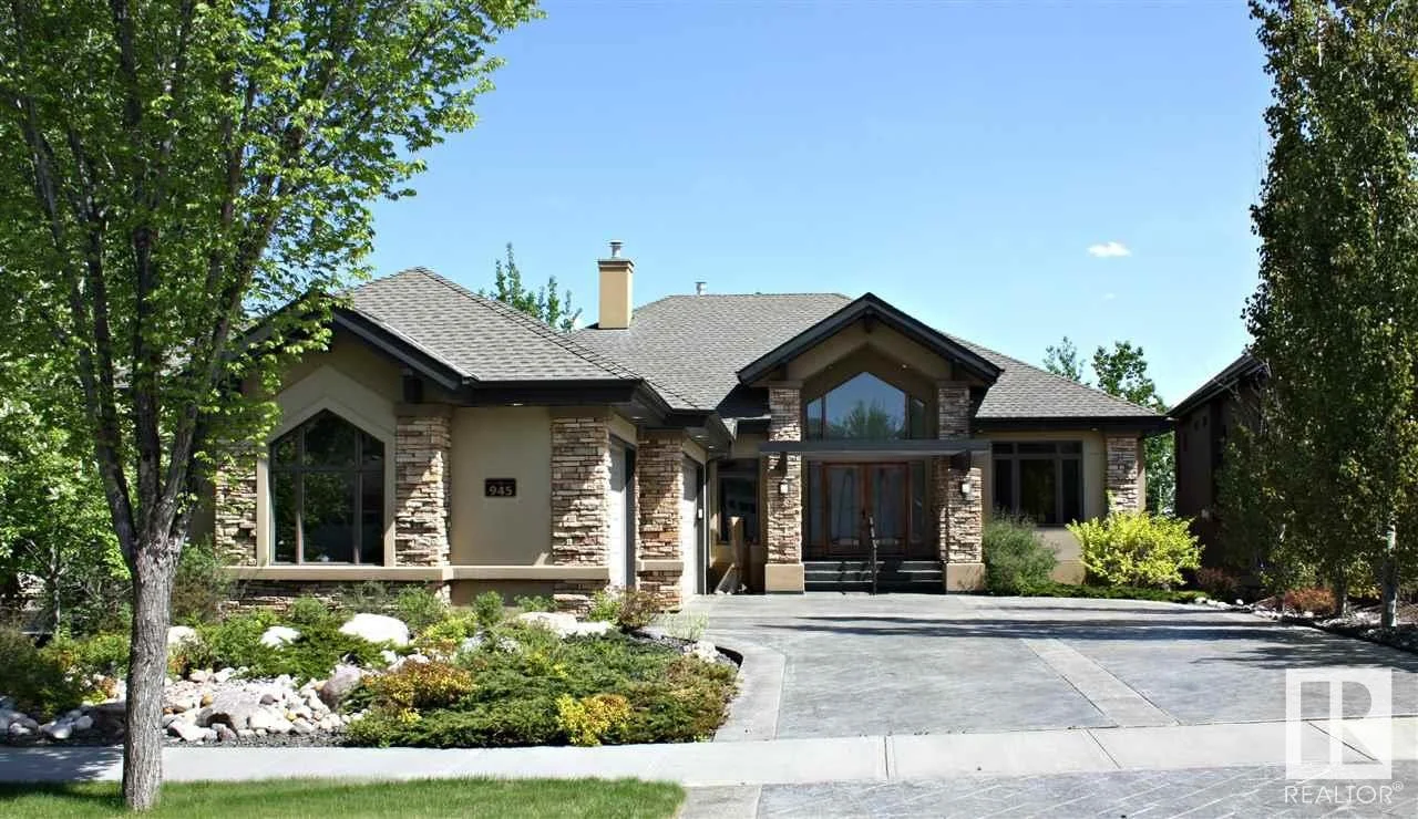 Modern house with stone facade, large front windows, and a paved driveway, surrounded by trees and landscaped garden.