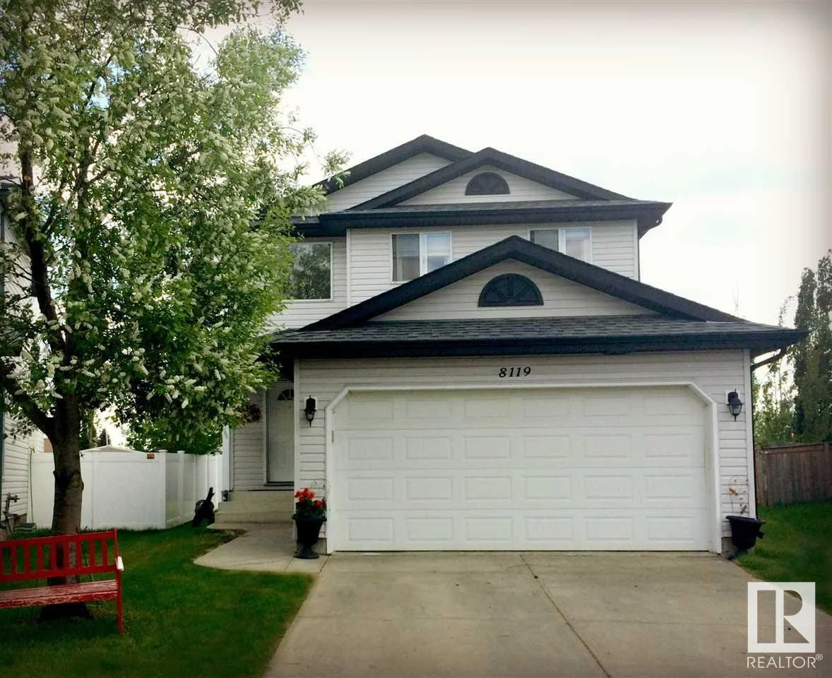 Two-story suburban house with white siding, black roof, attached garage, and front door with stairs, surrounded by green lawn, tree with white blossoms on the left, and a red bench on the left side of the yard.