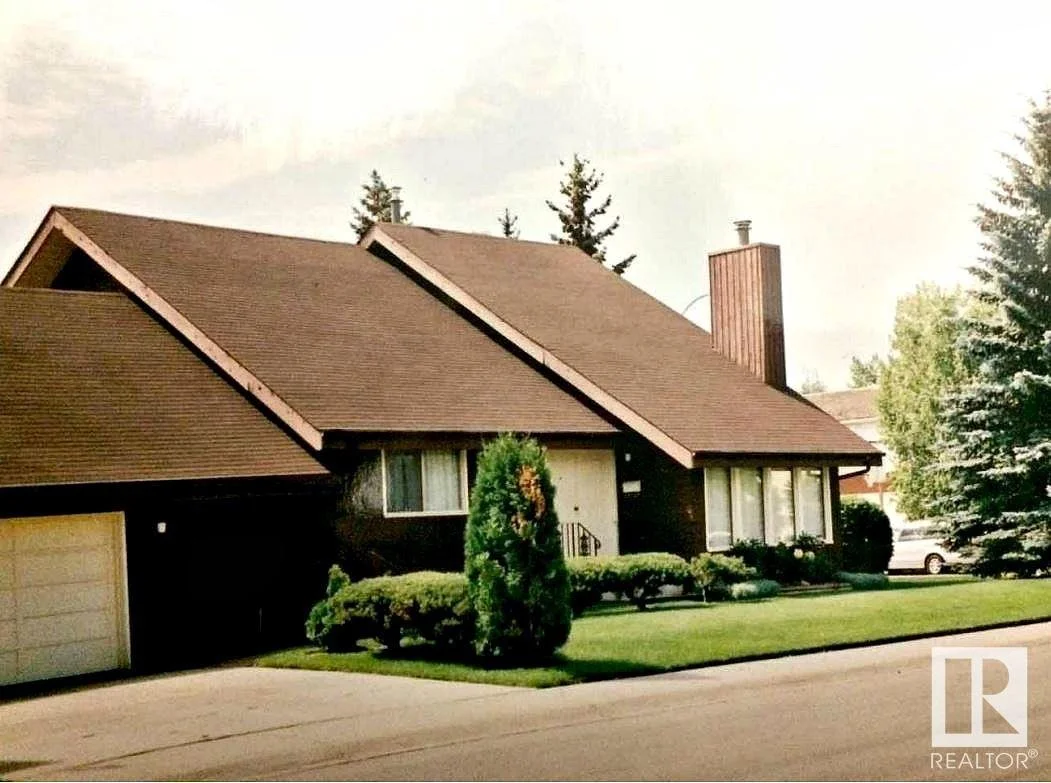 A suburban house with a steep gabled roof, a prominent chimney, a well-manicured lawn, and bushes along the front.