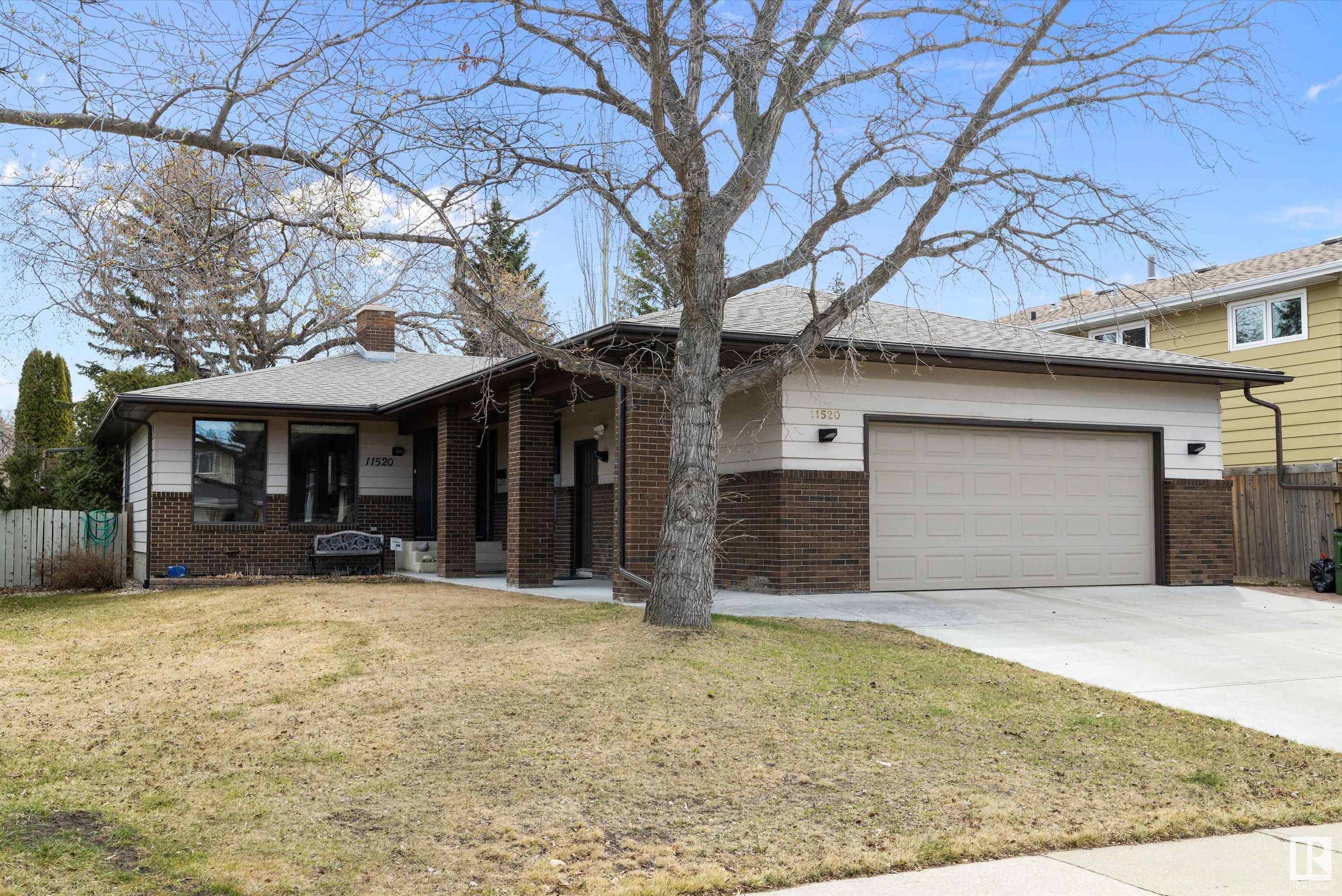 Front view of a single-story house with a brick and siding exterior, a large leafless tree in the front yard, and a driveway leading to an attached garage, under a clear blue sky.