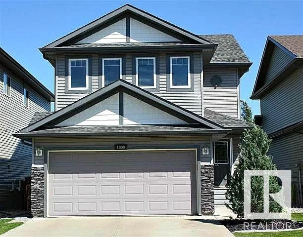 Two-story house with gray and white siding, a double garage, and a small front yard with a bush.