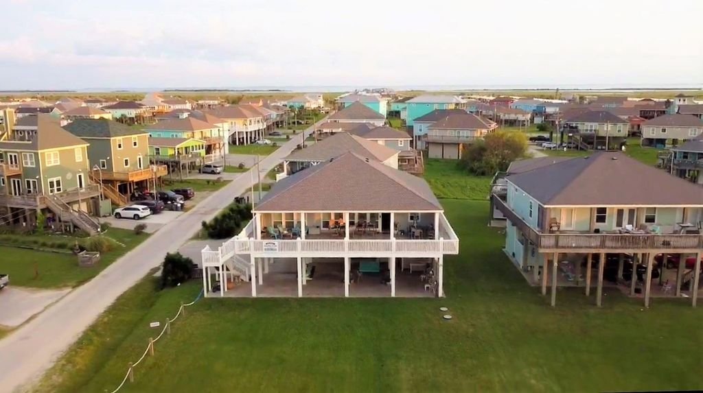Aerial view of a beachfront neighborhood with colorful houses on stilts, a paved road, and a grassy area in the foreground.