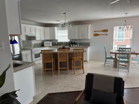 Open kitchen and dining area with white cabinets, a kitchen island with four chairs, a small dining table with four chairs, large windows, and modern decor.