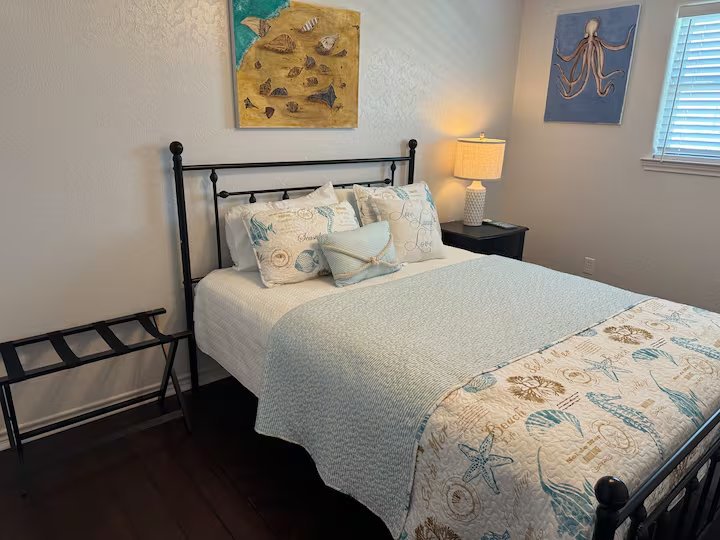 A bedroom with a metal bed frame, ocean-themed bedding, and decorative pillows. Two wall-mounted marine art pieces and a bedside lamp on a dark nightstand.