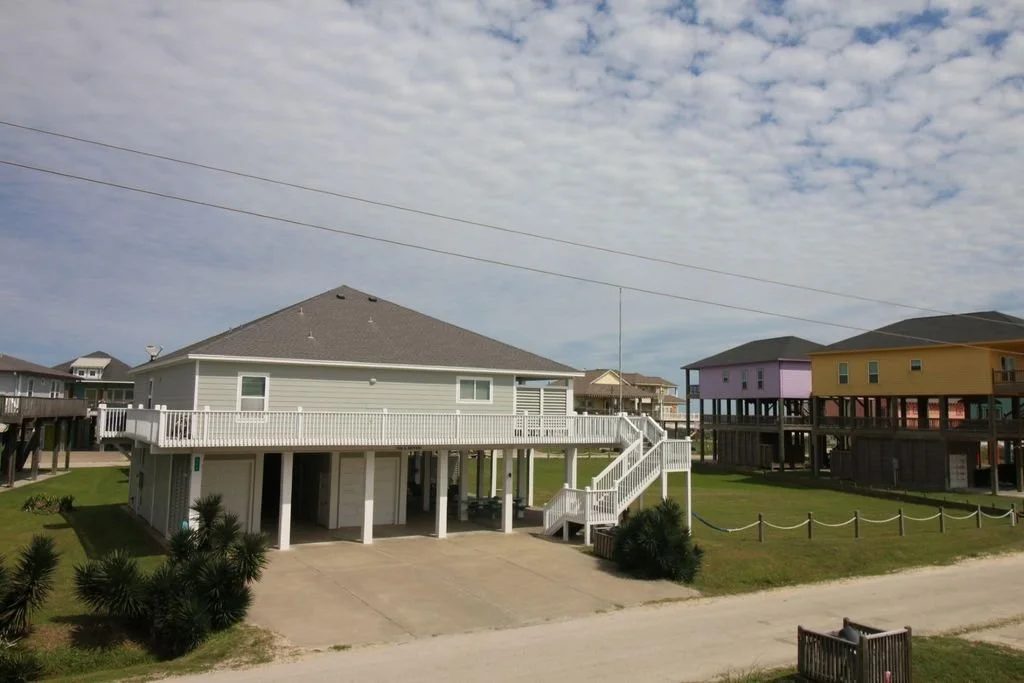 Colorful elevated beach houses on stilts with a blue sky and scattered clouds, some houses with decks and stairs, surrounded by green grass and a paved driveway.