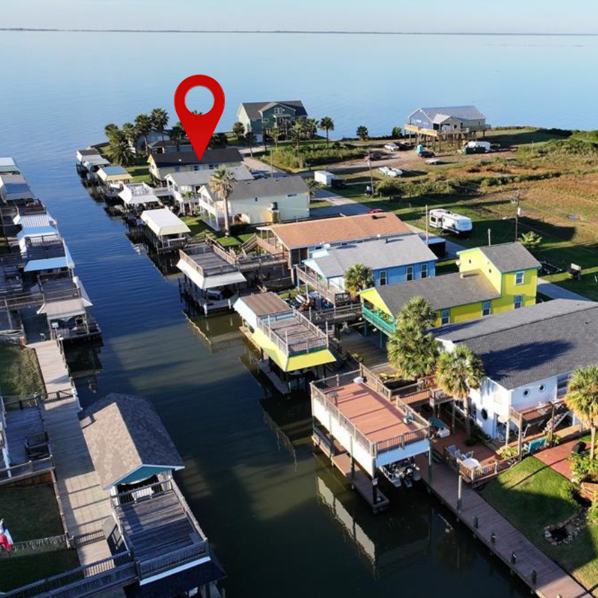 Aerial view of waterfront houses on stilts along a canal, with boats docked at their slips, and a body of water in the background.