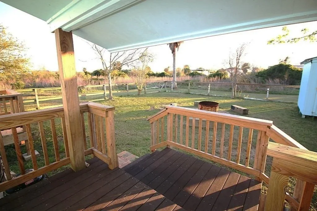 View from a wooden porch looking out onto a grassy backyard with a fence, trees, and clear sky during sunset.