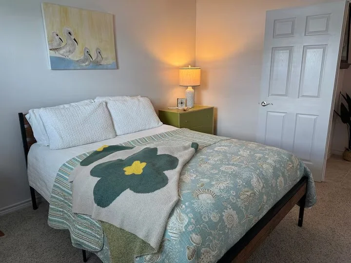 A tidy bedroom with a bed dressed in white and patterned bedding, a green nightstand with a lamp, and artwork of four ducks above the bed.