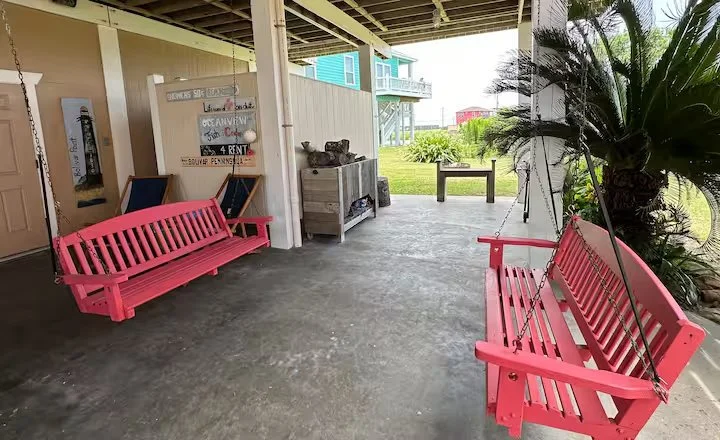 Covered porch area with pink painted wooden benches, a small storage shelf, a signboard, and greenery in the background.