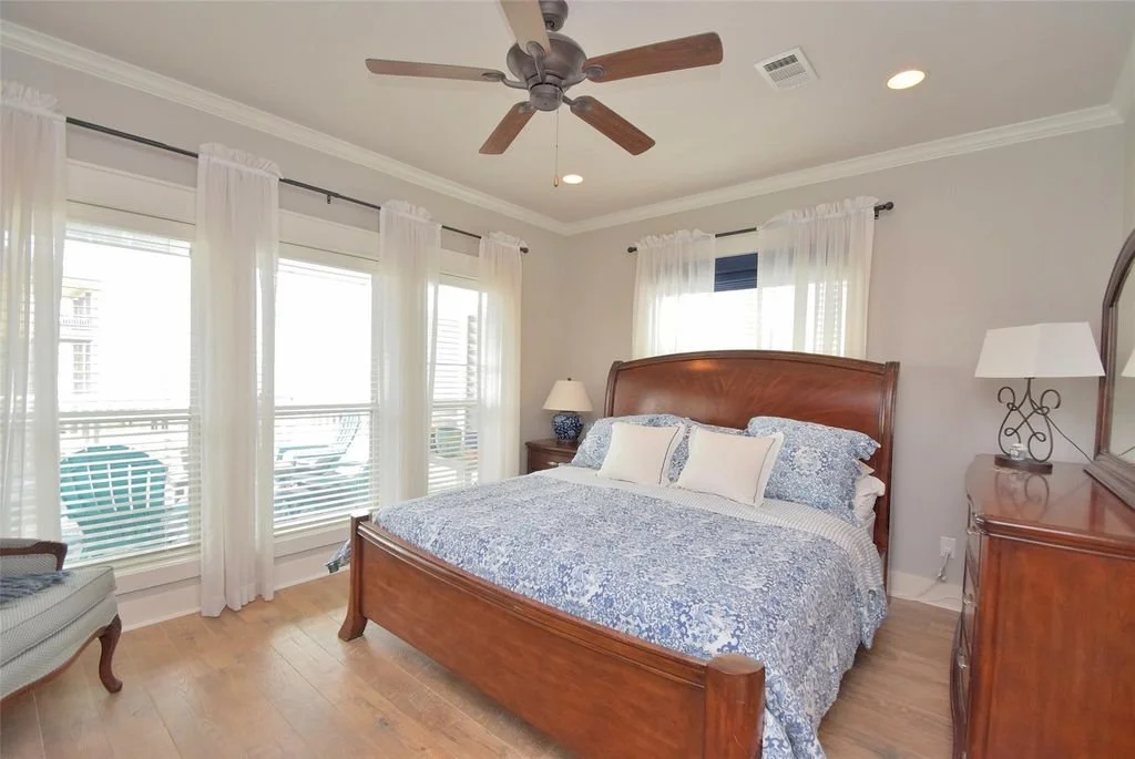 A bedroom with a large wooden bed, blue and white bedding, multiple pillows, a wooden dresser with a lamp, a side table with a lamp, and three windows with white curtains, light hardwood floors, and a ceiling fan.