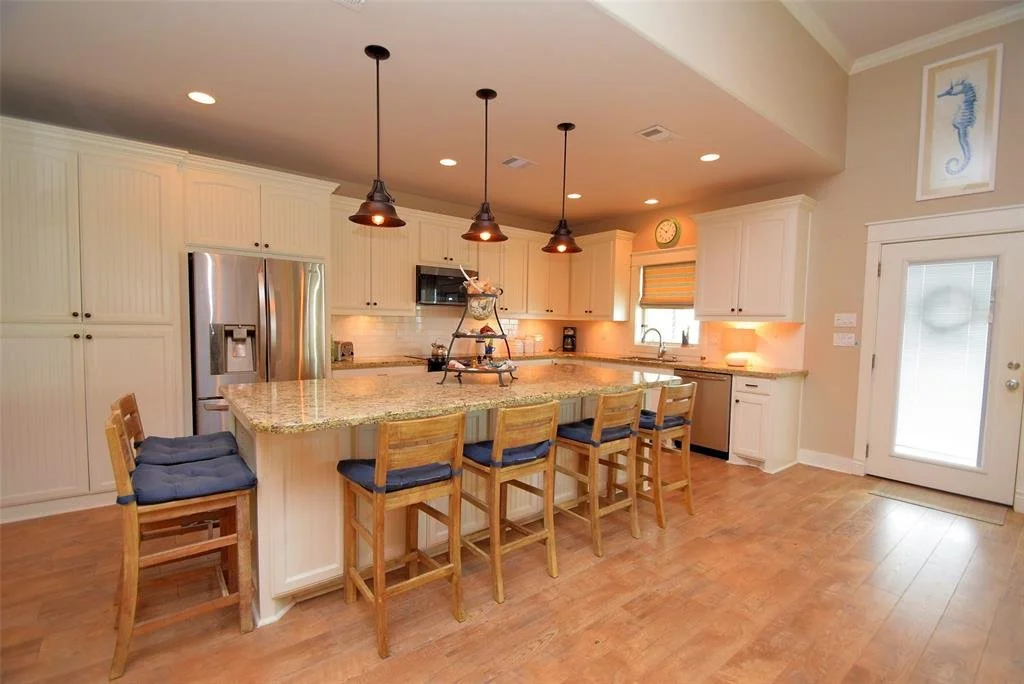 Bright kitchen with white cabinets, granite countertop island with four wooden chairs, stainless steel refrigerator, and hanging pendant lights.