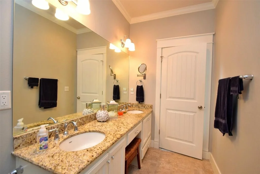 Bathroom with beige granite countertop, double sink, mirror, black towels, small round mirror, decorative jars, and soap dispenser.