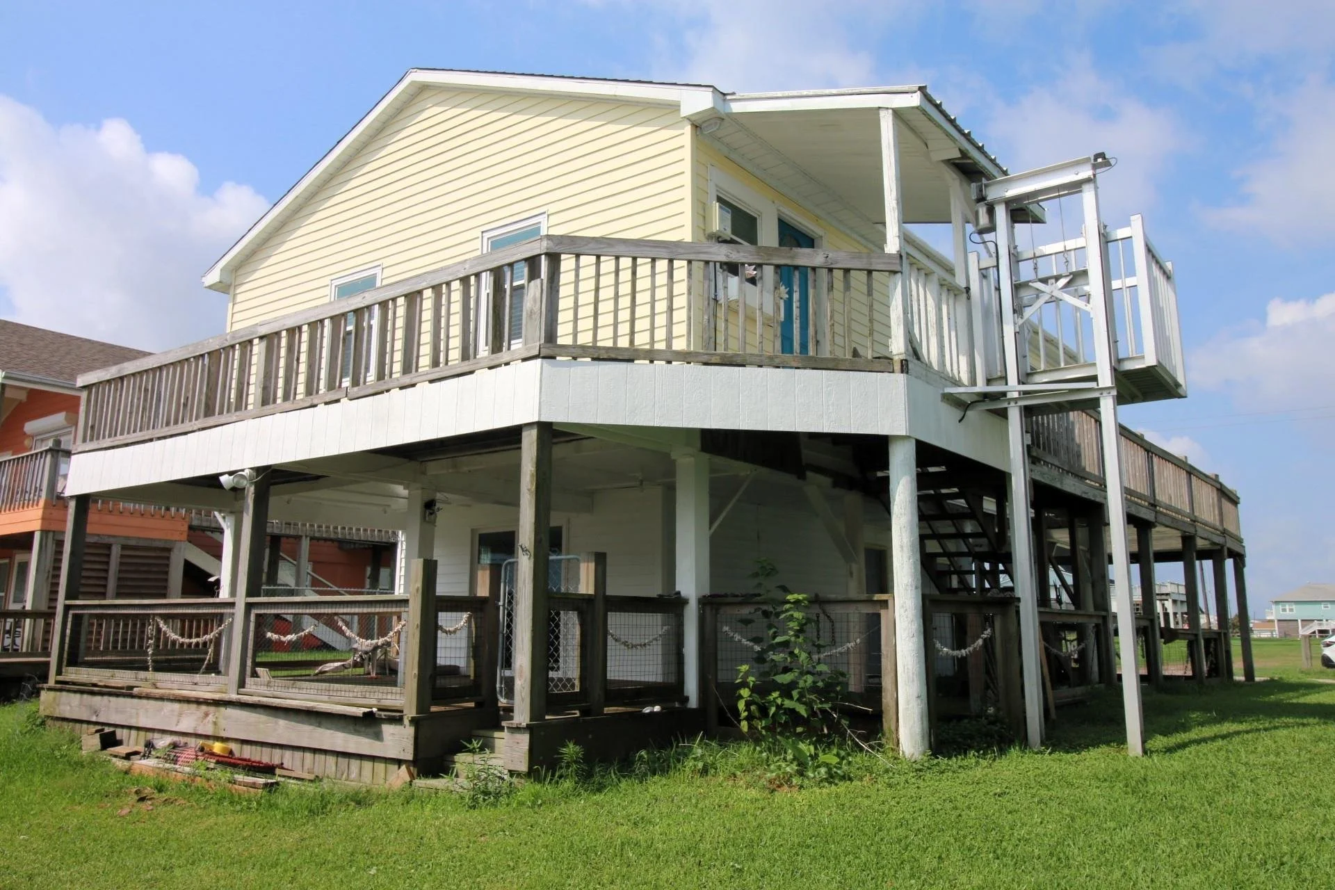 Beach house with yellow siding, white trim, and a wraparound deck on stilts with stairs leading down to the grassy yard; sky with some clouds.