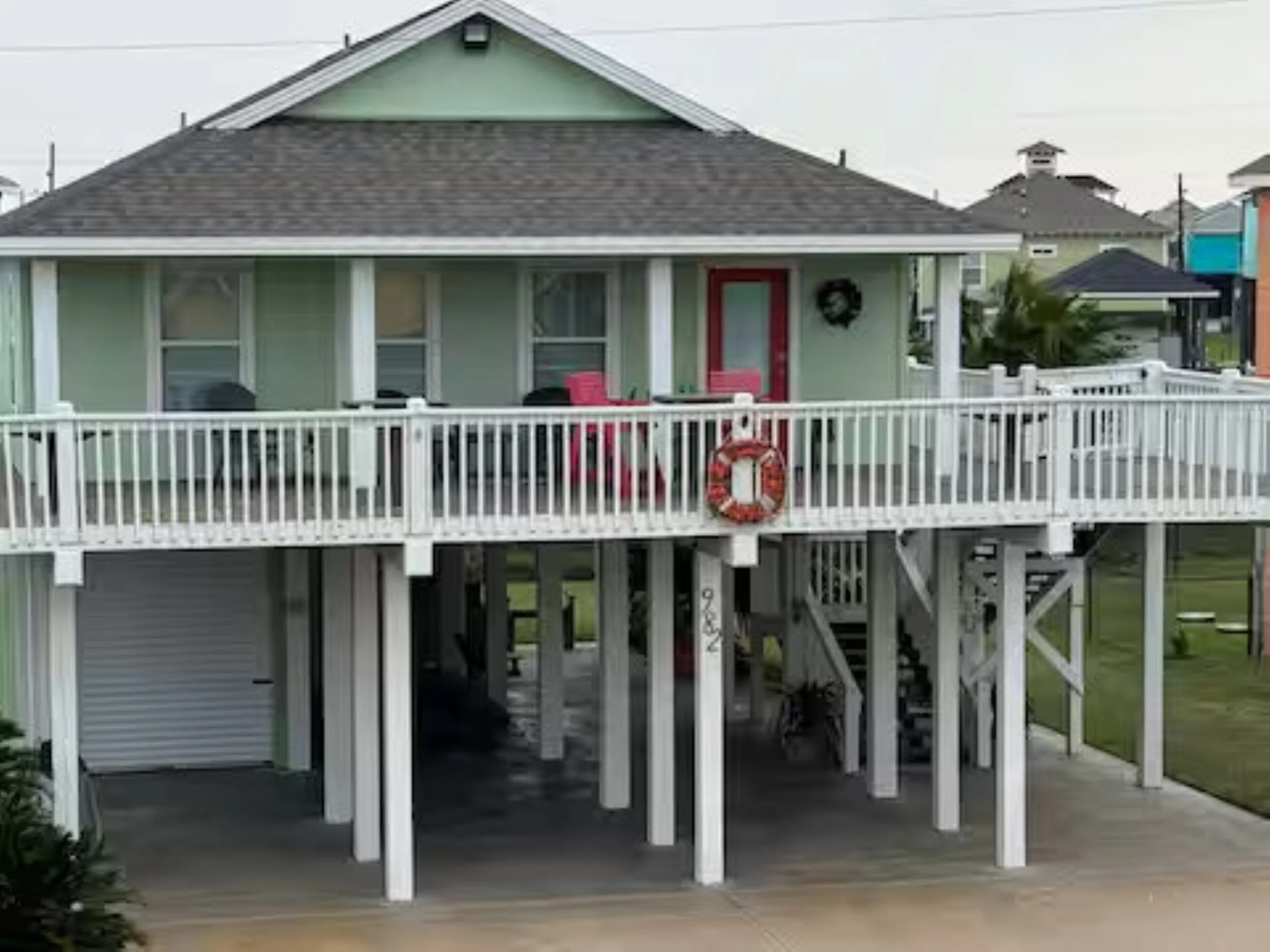 A two-story house with a porch, a red door, and a Life Buoy ring hanging on the wall. The house is painted light green with white trim and has a brown shingle roof. The house has a white railing around the porch and a staircase leading down to the sidewalk. There are neighboring houses visible in the background.