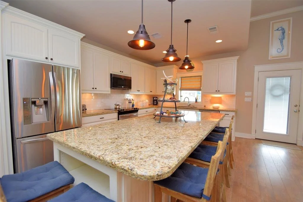 A modern kitchen with white cabinets, granite countertops, stainless steel refrigerator, microwave, oven, and a center island with seating. Pendant lights hang above the island. There is a small window, a door with a glass panel, and a seahorse artwo