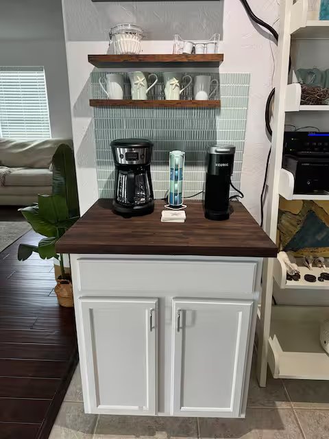 Coffee station with two coffee makers, a water filter, and cups on a wooden countertop against a tiled wall with shelves holding glasses and mugs.