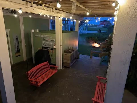 A cozy outdoor patio area with string lights, two pink benches, a small fire pit in the background, and a green wall with a mailbox and some decorative items.