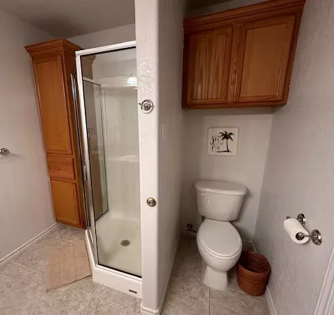 Bathroom with a corner shower enclosure, a toilet, wooden cabinets above and beside the toilet, a wall decoration with a palm tree, a wastebasket, and a roll of toilet paper.