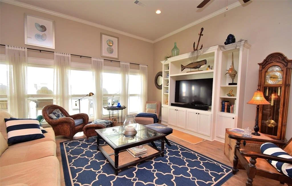 Living room with a beige sofa, wicker armchair, and a blue area rug. Large windows with sheer curtains, white built-in entertainment center with TV, nautical decorations, and a grandfather clock.